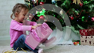 Little girl openning gift box near Christmas tree. Child tearing paper off gift