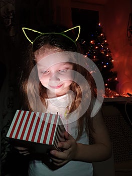 a little girl opens a box with a gift and is very happy with a surprise on Christmas evening near the New Year tree