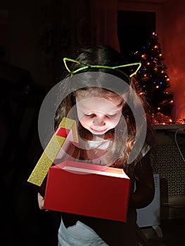 a little girl opens a box with a gift and is very happy with a surprise on Christmas evening near the New Year tree