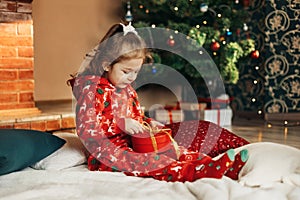 A little girl opens a Christmas present near the Christmas tree. Christmas time, gifts, festive mood. The concept of