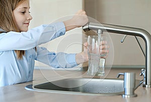 Little girl opens a water tap with her hand holding a transparent glass. Kitchen faucet. Glass of clean water. Filling cup