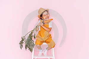 Little girl in an orange jumpsuit is sitting on stepladder. Child holding carrot in front of pink background