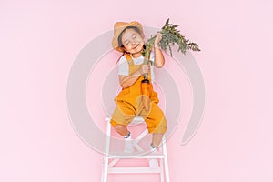 Little girl in an orange jumpsuit is sitting on stepladder. Child holding carrot in front of pink background