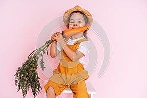 Little girl in an orange jumpsuit is sitting on stepladder. Child holding carrot in front of pink background