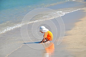 A little girl in an orange swimsuit sits on the beach on a sunny day.