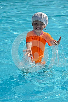 A little girl in an orange swimsuit is splashing in the pool on a sunny day.