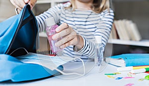 Little girl packing blue backpack in kids`s room. Face mask, bottle of sanitizer, stationery, pens, multicolored markers,