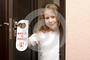 A little girl with painted lips peeks out from behind a door that says Do Not Disturb Make-up in Progress