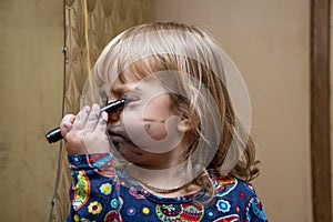 A little girl paints her face with a felt-tip pen in front of a mirror, a young makeup artist. Concept: childlike spontaneity
