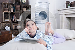 Little girl in pajamas lying on bed with digital tablet