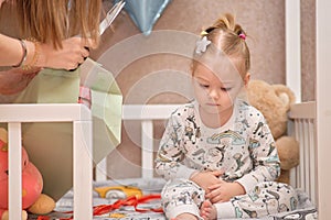 A little girl in pajamas receives birthday gifts while sitting in a white baby cot. Balloons in the background.