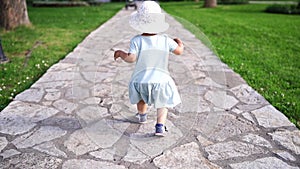 Little girl in a panama hat walks along a tiled path in the garden, staggering and balancing with her arms