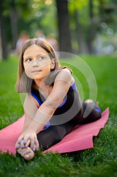 A little girl in the park is doing yoga stretching leaning on her leg. A girl is sitting on a twine