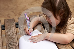 A little girl passionately draws on a piece of paper spread out on a bench in a street playground. Copy space.