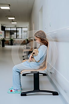 Little girl, patient, sitting alone at hospital corridor with her teddy bear.