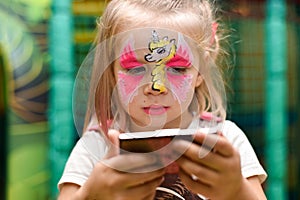 A little girl with a pattern of a unicorn on her face looks in the mirror