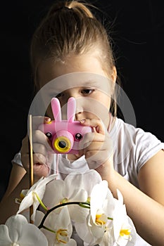 Little girl photographs a white orchid flower with a pink camera in the studio on a dark background