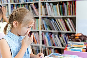 Little girl picking her nose,reading book with fairy tales in children`s library. Special reading kids room. Many shelves with