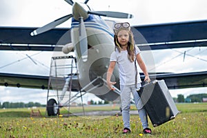 A little girl in a pilot`s costume carries a retro suitcase and walks along the airfield. A child in a hat and glasses