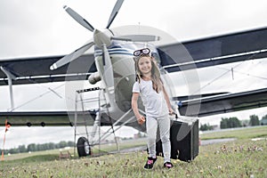 A little girl in a pilot`s costume carries a retro suitcase and walks along the airfield. A child in a hat and glasses