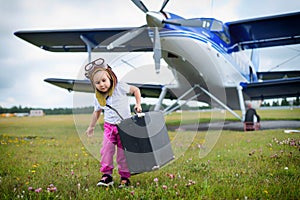 A little girl in a pilot`s costume carries a retro suitcase and walks along the airfield. A child in a hat and glasses