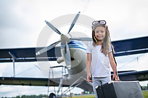 A little girl in a pilot`s costume carries a retro suitcase and walks along the airfield. A child in a hat and glasses