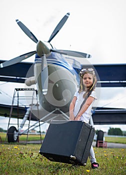 A little girl in a pilot`s costume carries a retro suitcase and walks along the airfield. A child in a hat and glasses