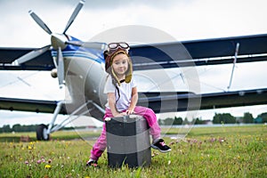 A little girl in a pilot`s costume carries a retro suitcase and walks along the airfield. A child in a hat and glasses