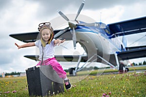A little girl in a pilot`s costume carries a retro suitcase and walks along the airfield. A child in a hat and glasses