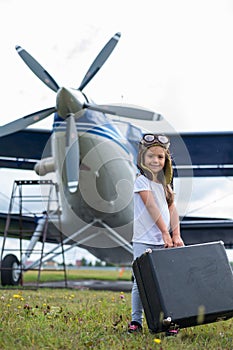 A little girl in a pilot& x27;s costume carries a retro suitcase and walks along the airfield. A child in a hat and glasses