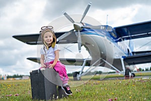 A little girl in a pilot's costume carries a retro suitcase and walks along the airfield. A child in a hat and glasses