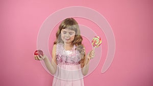 Little girl on a pink background holding a red apple and candy.