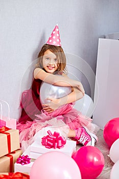 Little girl in pink dress and birthday hat sitting at the floor with many present gift box, balloons