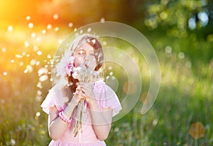 Little girl in a pink dress blowing a bouquet of dandelions in the rays of a bright sun