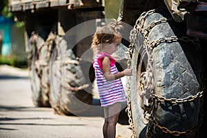 A little girl in a pink dress next to the big wheels of a car in chains.