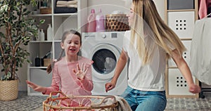 A little girl in a pink dress and pigtails sits with her mother on the laundry room floor with a wicker basket and sorts