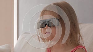 A little girl in a pink dress sitting on the couch attentively and emotionally watching TV in 3D glasses