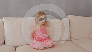 A little girl in a pink dress sitting on the couch attentively and emotionally watching TV in 3D glasses