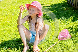 A little girl in a pink hat sits on the fresh grass and holds in her hands a butterfly caught in a butterfly net.