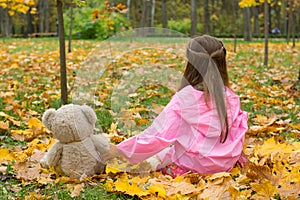 A little girl in a pink raincoat is holding a teddy bear behind her paw in the autumn.