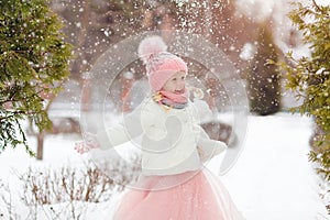 Little girl in a pink skirt in winter smiles in the park and throws up the snow