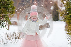 Little girl in a pink skirt in winter smiles in the park and throws up the snow