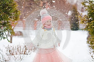 Little girl in a pink skirt in winter smiles in the park and throws up the snow