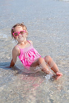 Little girl in pink swimsuit and sunglasses sitting in the sea posing at camera. Happy, smiling. Summer vacation.