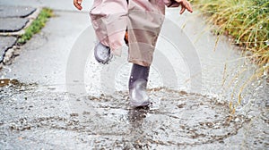 Little girl in pink waterproof raincoat, purple rubber boots funny jumps through puddles on street road in rainy day