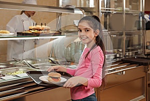 Little girl with plastic tray and burger near serving line in canteen