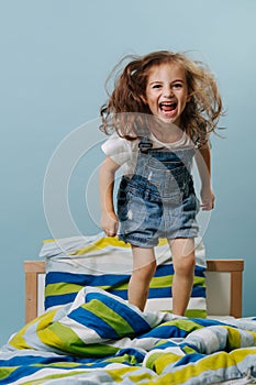 Little girl is playing actively, jumping in bed over blue background