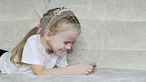 Little girl is playing on her smartphone and laughing while lying on the couch