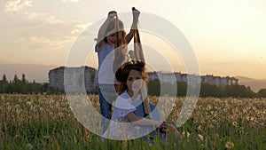 Little girl playing with long hair of her sister with guitar sitting on dandelion grass field in suburban on sunset