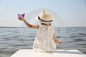 Little girl playing with paper boat on wooden pier near river, back view